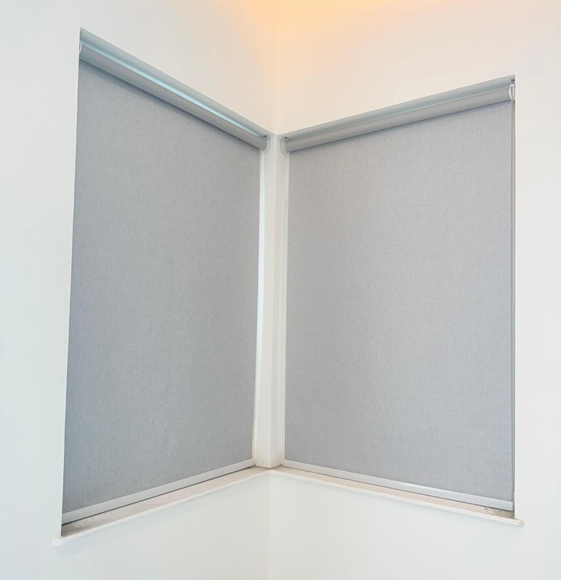 Two grey roller blinds meeting at a corner window