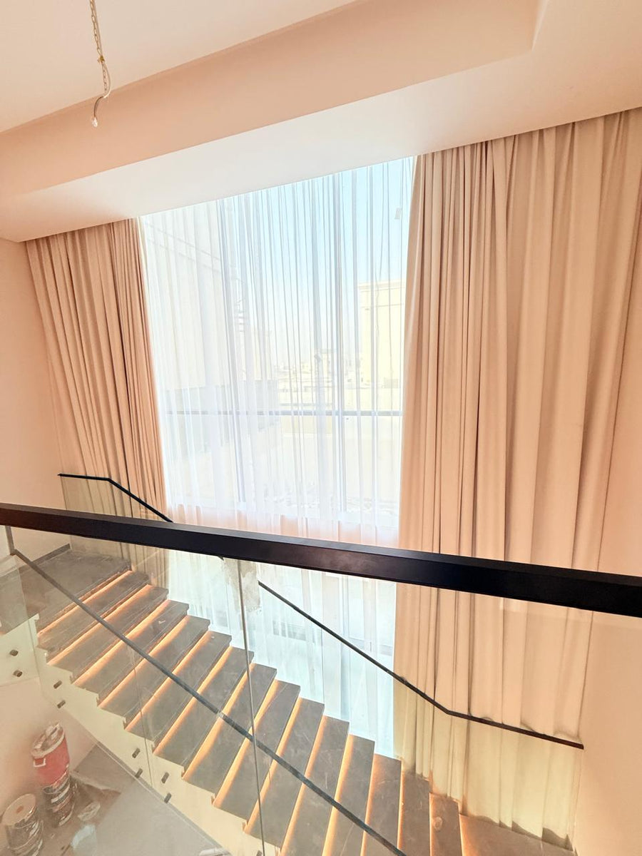 Blush curtains and white sheer at a double-height stairwell window above a timber staircase