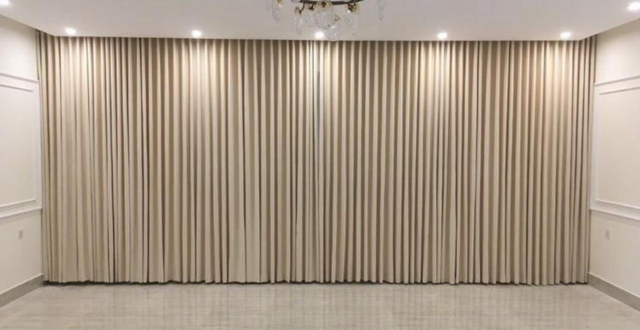 Taupe wave curtains drawn fully closed across an entire panelled wall in an empty room lit only by ceiling lights