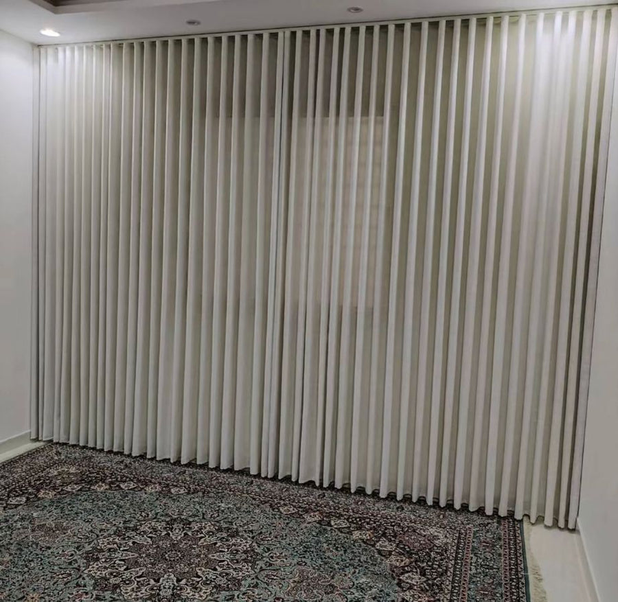Off-white wave curtains drawn closed around a corner run, daylight showing faintly through the fabric, above a patterned rug in an empty room